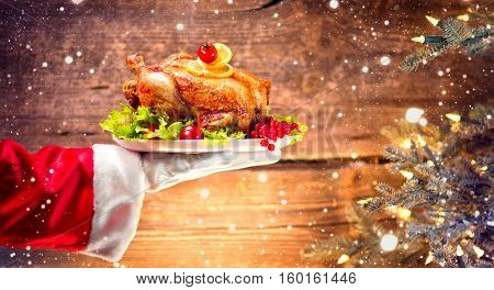 Christmas Holiday dinner. Santa Claus hand holding roasted Chicken. Christmas and New Year food concept, over rural wooden background and Decorated Christmas tree with lights.