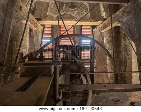 Picture of the rusty mechanism in the wooden belfry of the church. The wall of the belfry made of timber painted wih red colour. The sunlight shining feebly through the slit between red planks.