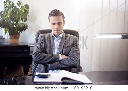 Male adult man in a suit sitting on a chair behind the desk in a office, looking down to notepad with calculator, arms crossed, indoor