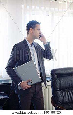 Male adult businessman in a suit talking on a cellphone and holding file standing in a office next to the window