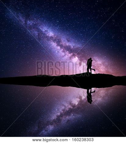 Milky Way. Night landscape with silhouettes of hugging and kissing man and woman on the mountain near the river with sky reflection in water. Starry sky. Lovers. Couple. Purple milky way with people