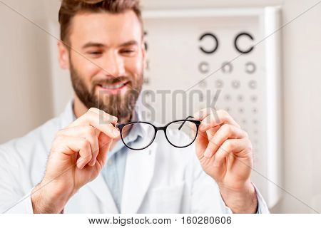 Handsome ophthalmologist looking at the glasses in front of the eye chart in the cabinet. Image with small deph of field focused on hands and glasses