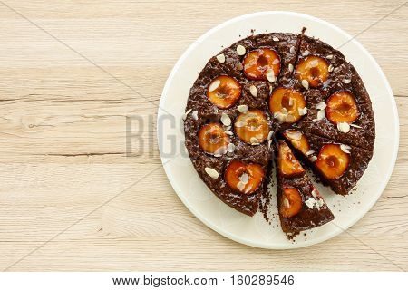 Closeup of a whole homemade Sticky Chocolate Plum Cake on a porcelain plate with one piece of cake cut. Isolated on light brown wooden background with lots of copy space.
