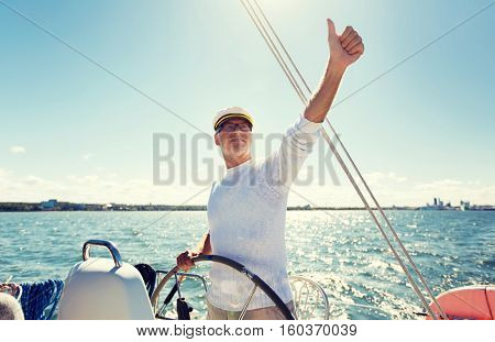 sailing, age, tourism, travel and people concept - happy senior man in captain hat on steering wheel and showing thumbs up sail boat or yacht floating in sea