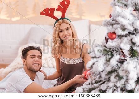 Beautiful Christmas tree. Young delighted bearded man holding a Christmas decoration and being in a great mood while helping his girlfriend to decorate the Christmas tree