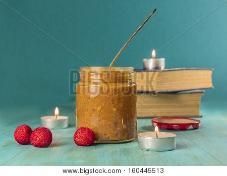A photo of a jar of madrono, wild strawberry, fruit butter, with a spoon, madrone berries, candles and old books. Selective focus