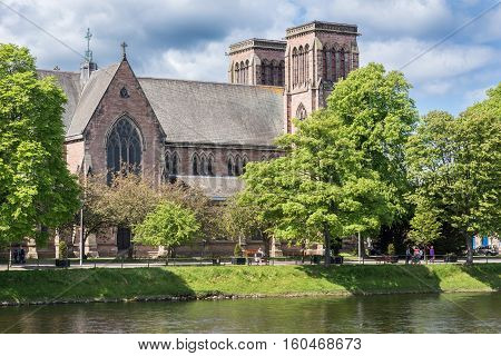 Inverness Scotland - June 1 2012: Brown stone Saint Andrews Cathedral show side roof and the two towers. White cloudy sky. Seen from across Ness River. Green trees.