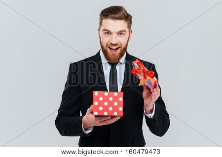 Happy bearded business man in suit opening gift and looking at camera. Isolated gray background