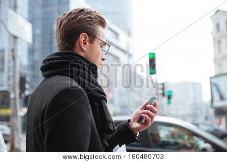 Back view of business man in glasses and warm clothes standing on the street and looking at phone