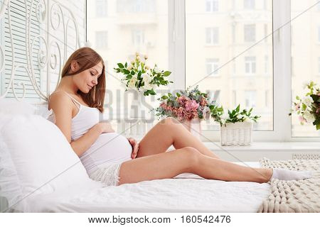 Mid shot of pregnant woman lying on bed and touching her belly enjoying the cheerfulness of the early morning. Holding her belly while sitting on the bed