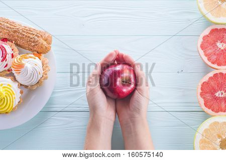 Hands of a young woman holding a red apple. Woman making a choice between sweets and fruits made a choice in favor of fruits and holding an apple. Weight Loss. Unhealthy vs healthy food top view.