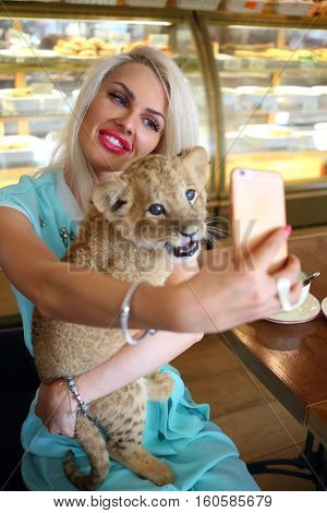 Beautiful woman holds funny calf of lion at table in cafe and makes selfie