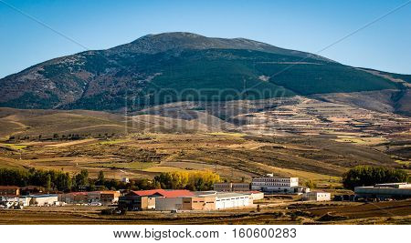 View of mountain with community buildings & homes in foreground in rural Spain.  Evidence of deforestation seen in terraced land as farm fields along straight edge of trees ascending mountainside.