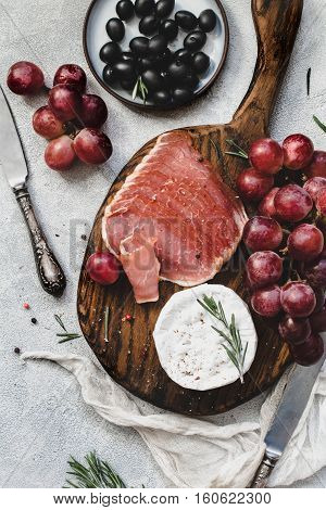 Assorted italian antipasti snacks for a dinner with wine or party on wooden cutting board. Red grapes, fresh camambert cheese, prosciutto and black olives. Top view, desaturated colors