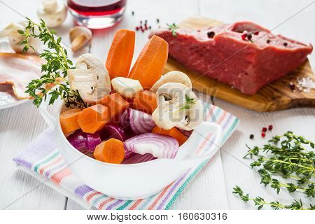 Ingredients for Boeuf Bourguignon carrot onion mushrooms bacon wine beef garlic thyme. White bowl wooden table.