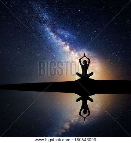 Milky Way. Silhouette of a sitting woman practicing yoga on the hill near the lake with sky reflection in water. Landscape with meditating girl Night starry sky and milky way with yellow light. Space