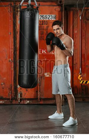 Photo of young strong boxer posing in a gym with punchbag. Looking at camera.