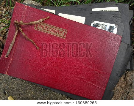 An old red leather family photo album with frayed cording and the word, Photographs, printed in gold opens slightly to show black paper pages with black and white photos.