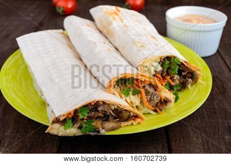 Shawarma sandwich - fresh roll of thin lavash (pita bread) filled with grilled meat mushrooms cheese cabbage carrots sauce green. Traditional Eastern snack. On a dark wooden background.