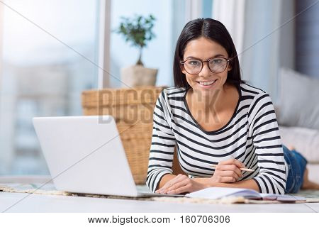 Enjoyable process of creating a project. Positive delighted smiling woman lying on the floor at home in front of the laptop while making notes in her notebook and working on the project