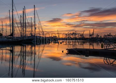 Twilight at harbor sunrises silhouettes of docked sailboats and cargo port cranes red orange colors in the sky and water reflection horizon