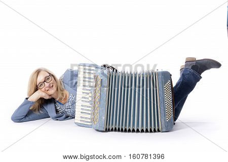 blond woman in blue with glasses lies on floor behind accordeon and smiles in studio with white background