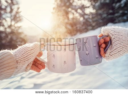 Man and woman hands in knitting mittens with cups of hot tea on winter forest glade