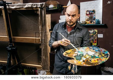 Artist looks at his painting with a brush and palette in his hands Artistic process in workroom