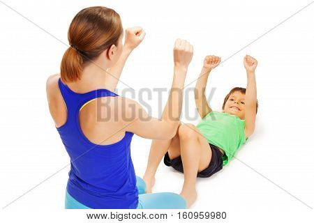 Smiling happy boy doing exercises laying flat on back and raising his hands up with female trainer, isolated on white