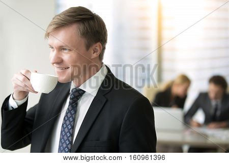 Young smiling businessman drinking tea or coffee, in modern office, dreaming of making fabulous income, look forward to holidays, overcame challenges and having a rest, shift responsibility to others
