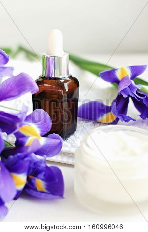 Floral essence, jar of facial cream, fresh lilac Iris flowers. Natural skincare cosmetic products. Vertical. Focus on dark glass dropper bottle.