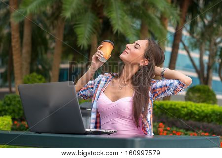 Young woman taking a break to relax from work on the laptop in park while holding a cup of coffee in one hand with her eyes closed and a smile of pleasure.