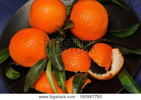 Ripe orange tangerines with green leaves in plate on black plate, top view. Low key. Concept of healthy nutrition, diet, detox