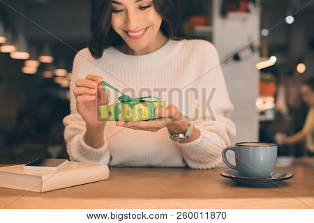 Smiling Young Woman Untying Gift Box At Table With Smartphone And Coffee Cup In Cafe