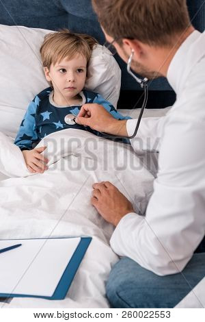 High Angle View Of Pediatrician Litening Childs Breath With Stethoscope While He Lying In Bed