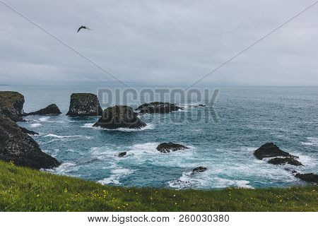 Dramatic Shot Of Rocks In Blue Ocean In Arnarstapi, Iceland With Green Meadow On Background