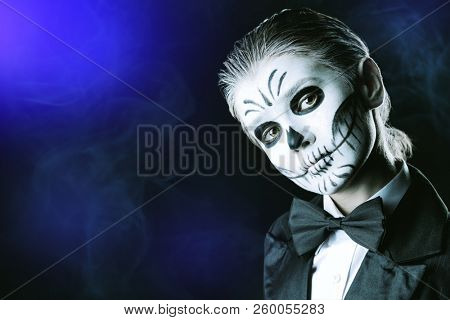 Portrait of a child boy with carnival make-up of a skeleton wearing elegant tail-coat. Studio shot over black background. Halloween. Dia de los muertos. The Day of the Dead.