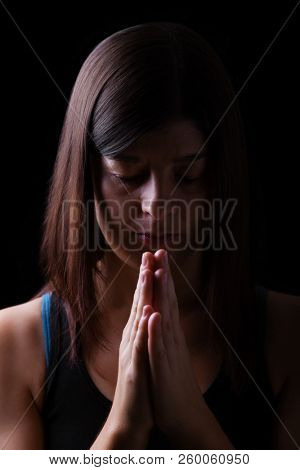 Faithful athletic woman praying, with hands folded in worship to god, head down and eyes closed in religious fervor, on black background. Concept for religion, faith, prayer and spirituality
