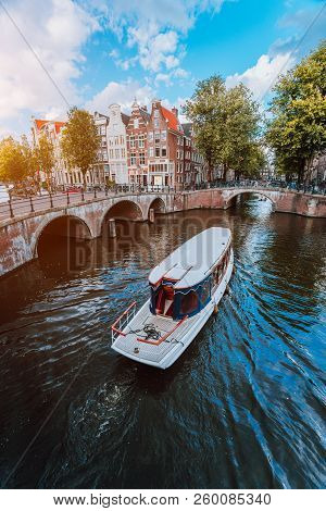 Tour Boat At Famous Dutch Canal On A Sunny Day, Traditional Dutch Bridges, Medieval Houses. Amsterda