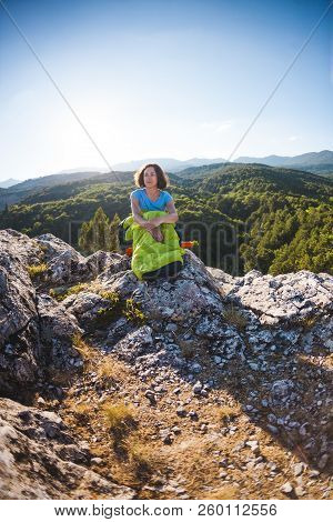 A Woman Is Sitting In A Sleeping Bag At Sunset. Camping On Top Of A Mountain. A Smiling Girl Rests A