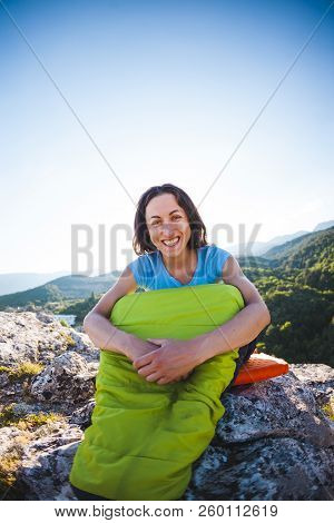 A Woman Is Sitting In A Sleeping Bag At Sunset. Camping On Top Of A Mountain. A Smiling Girl Rests A