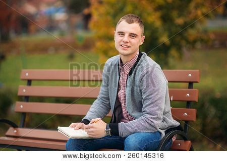 A Cute Student Sits On The Bench And Writes Down His Thoughts In His Notebool Using A Pensil. Study 
