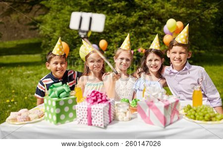 holidays, childhood and technology concept - happy kids taking picture by selfie stick on birthday party at summer garden