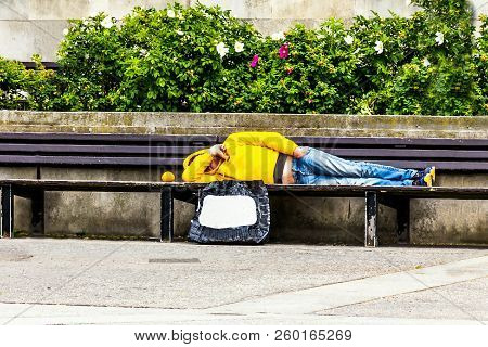 Unrecognizable Unemployed In A Yellow Jacket With A Hood And Blue Jeans Lies On A Park Bench, Coveri