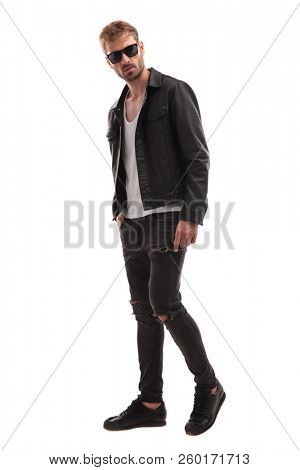 side view of young blond man wearing sunglasses and leather jacket stepping to side on white background, full body picture