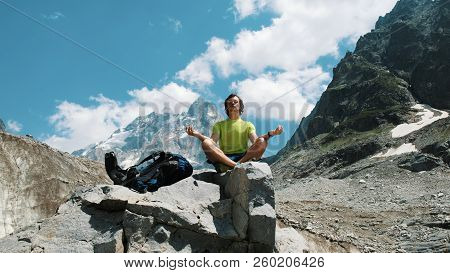 Tourist Man Hiker Meditating In The Mountains Sitting On A Stone, Relaxing And Pacifying In The Wild