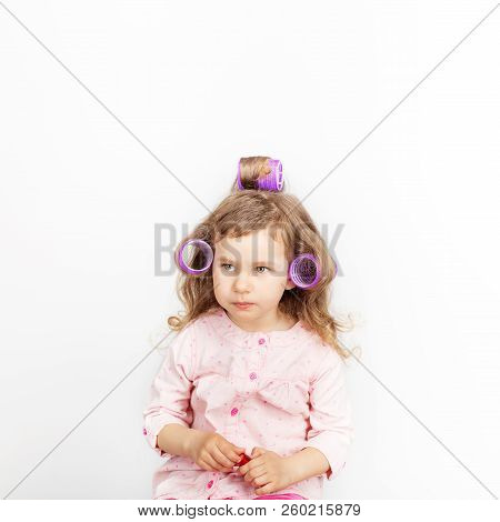 Beautiful Little Girl With Curlers On Her Head Looking At Camera And Having Fun At Home