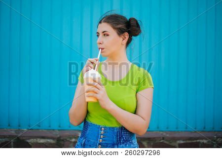 Portrait Of A Girl Against A Bright Blue Wall. Emotional Girl Holding A Plastic Glass With A Refresh