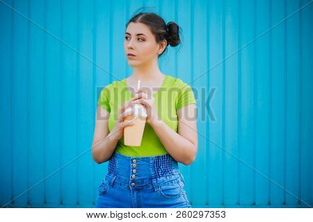 Portrait Of A Girl Against A Bright Blue Wall. Emotional Girl Holding A Plastic Glass With A Refresh