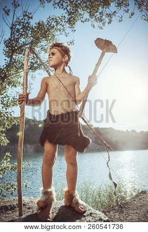 Caveman, Manly Man With Stone Axe And Bow Hunting Near River. Prehistoric Tribal Boy Outdoors On Nat
